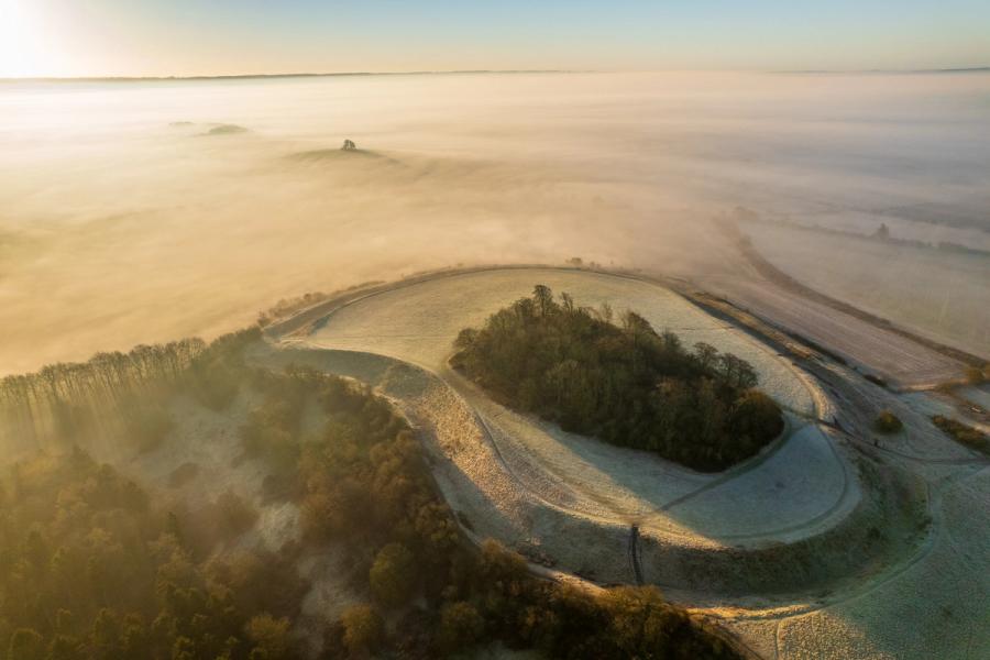 At first light on a late-winter morning, Brightwell Barrow near the Wittenham Clumps in Oxfordshire emerges frost-rimed from a temperature inversion above the Vale of White Horse, its concentric earthwork ramparts carving a near-perfect ellipse through the frosted grass. A sea of fog erases the surrounding vale while the tree-crowned knoll of the Wittenham Clumps floats at the horizon. The composition holds the rare alignment of prehistoric geometry, seasonal atmosphere, and aerial perspective that rarely arrives twice.