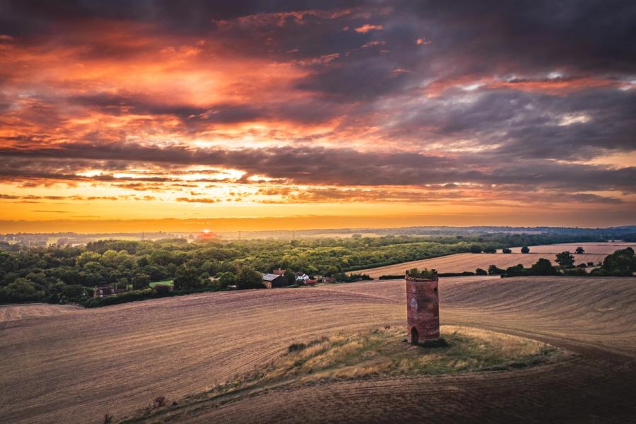 A ruined brick tower stands alone on a harvested hillside above the Thames Valley, south of Reading, as the sun drops below a distant tree line. Heavy cumulus catches the last orange light while mist gathers in the valley floor below. The composition uses the folly as a solitary anchor against a wide, dramatic sky.