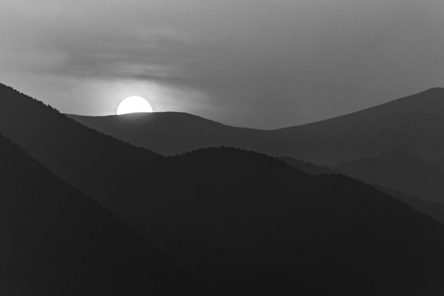A low sun sits precisely on a moorland ridgeline, its disc rendered luminous against overcast cloud while successive hill silhouettes recede into atmospheric haze. The tonal layering â from near-black foreground slopes through mid-grey ridges to a bright corona â is handled with considerable restraint. Captured late afternoon in early spring, the image reads as monochrome conversion rather than in-camera black-and-white.