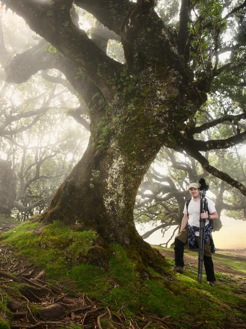 Christopher Harrison beside an ancient laurel tree in the Fanal Forest in Madeira, with his camera and tripod in hand, backed by misty trees.