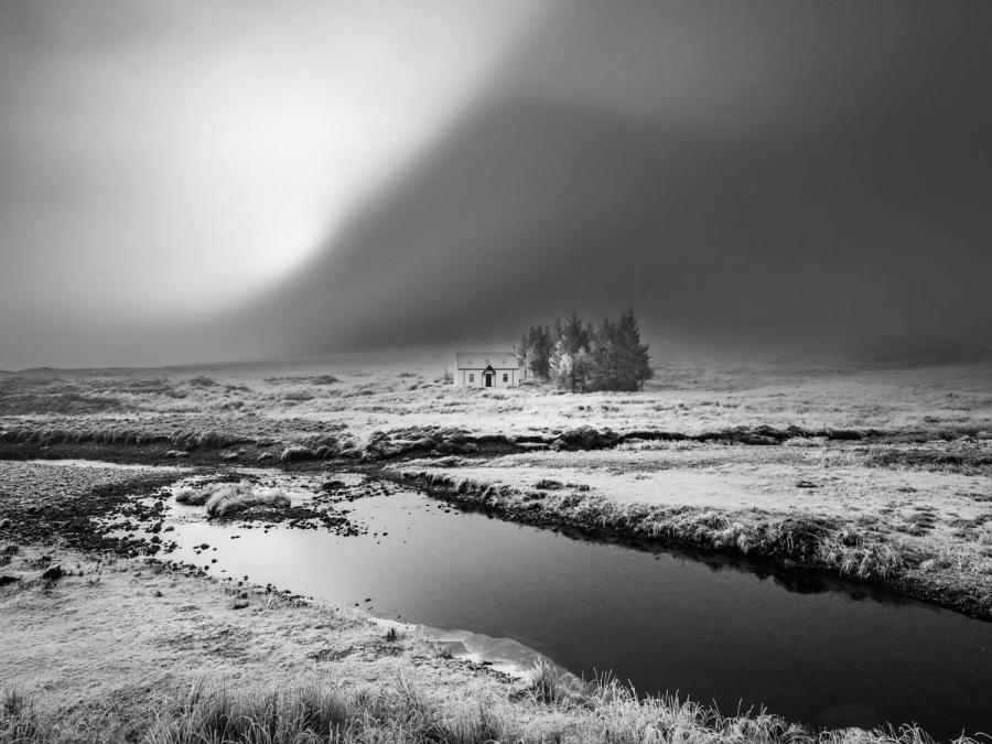A solitary farmhouse and small stand of conifers sit exposed on a frost-bleached moor as a storm front sweeps overhead, its dark mass bisected by a shaft of diffuse light. A shallow moorland stream curves through the foreground, its still surface reflecting the unsettled sky. The tonal contrast between the luminous cloud break and the advancing darkness gives the image its underlying tension.