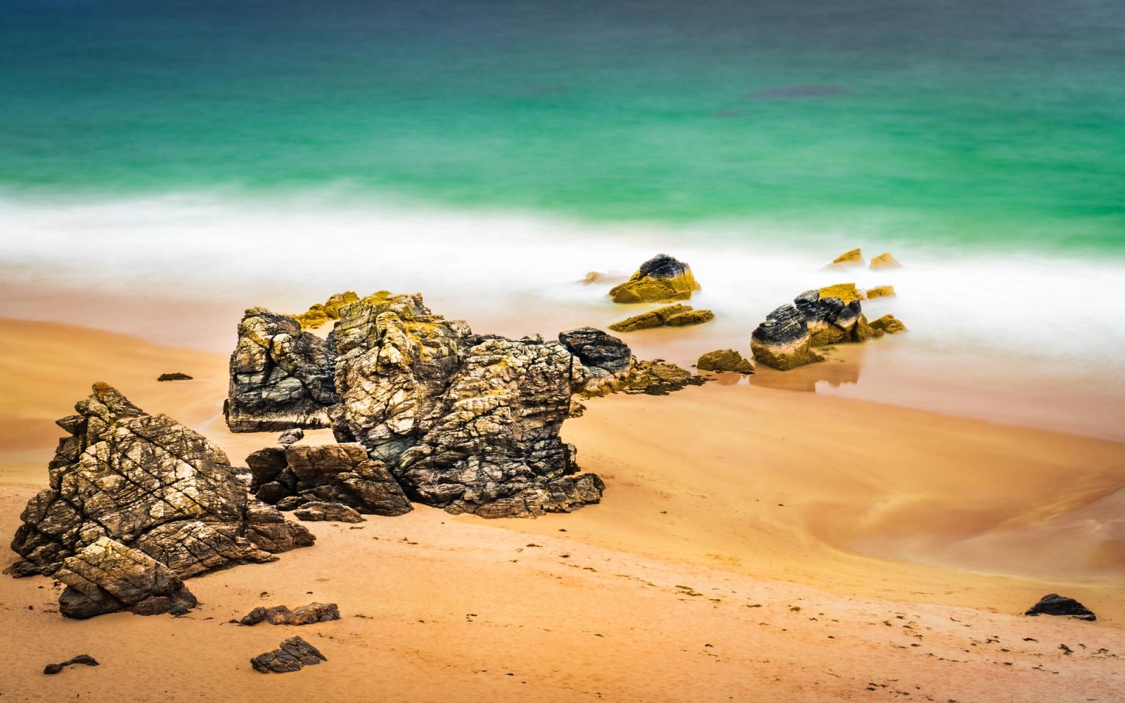 Fractured coastal rocks scatter across a sandy beach as long exposure reduces the incoming surf to a smooth, luminous wash. The vivid turquoise of the water contrasts with the warm ochre sand, the motion-blur lending an almost dreamlike stillness to an otherwise turbulent shoreline. Captured mid-afternoon in late spring.