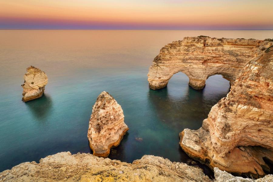 Twin sea arches cut through limestone headland as the last afterglow settles over a calm Atlantic. Long exposure has smoothed the water to a pale teal, drawing the eye through the archways to the open horizon. Isolated sea stacks punctuate the foreground cove.
