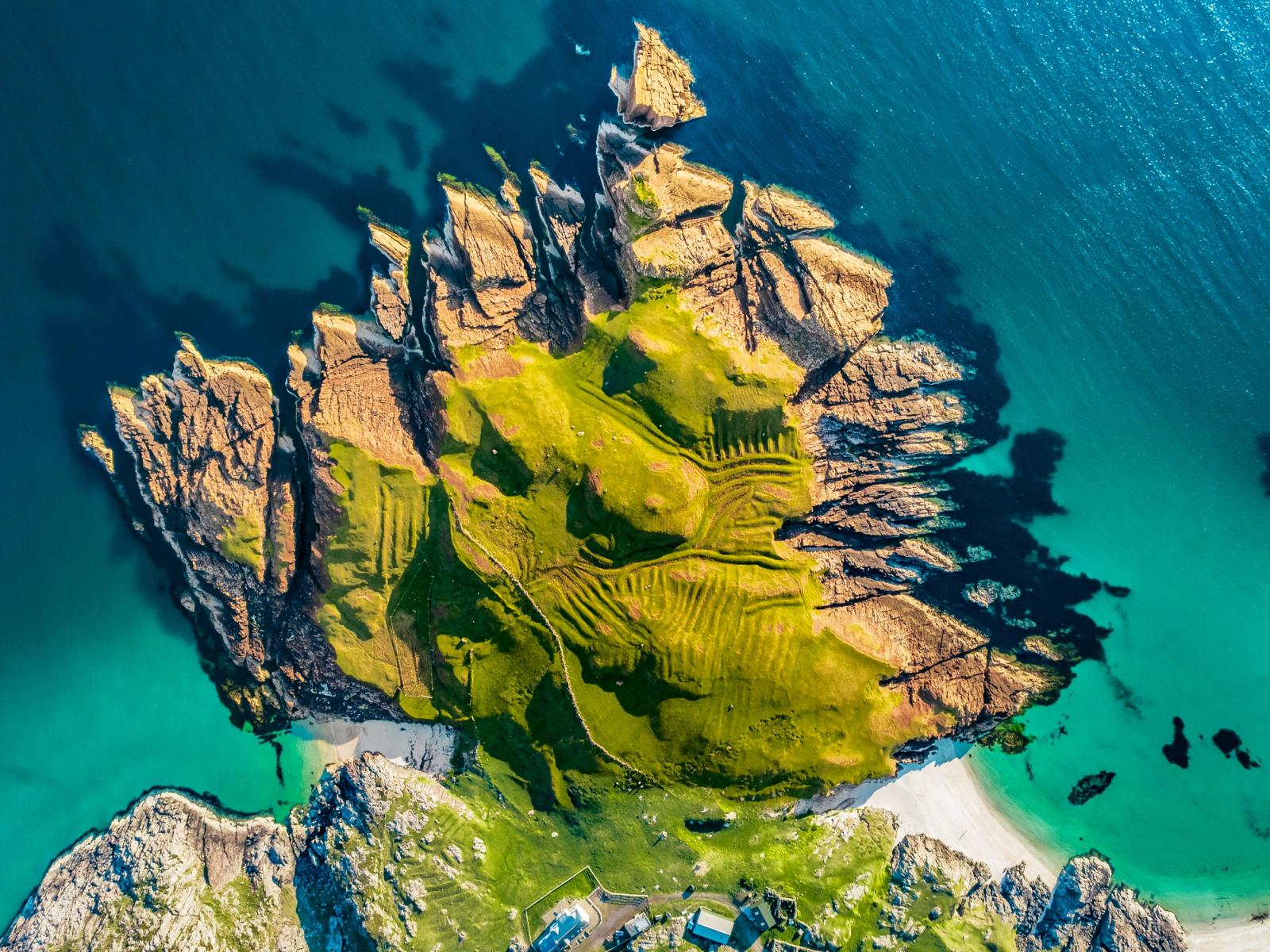 A tidal headland on the north-west Scottish coast, photographed from altitude in late evening light. Folded ancient gneiss strata radiate outward like fins from a grass-capped central plateau, while ancient lazybeds score the turf in parallel ridges. The surrounding sea shifts from deep indigo to shallow turquoise where sand lies beneath the surface.