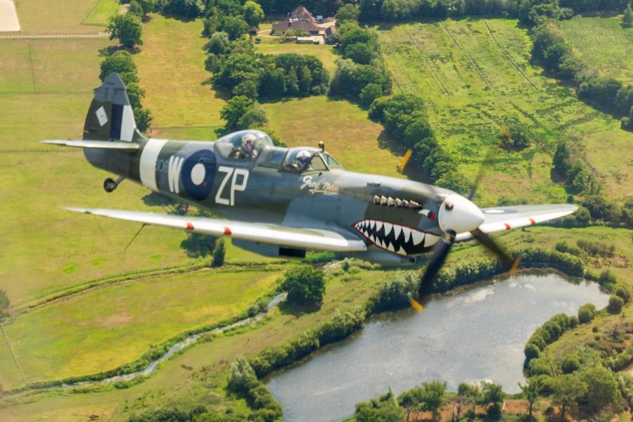 A two-seat Supermarine Spitfire, marked 'ZP-W' and named Grey Nurse with shark-mouth nose art, crosses a patchwork of summer fields and a small lake somewhere over lowland England. The air-to-air perspective places the aircraft in tight relationship with the rural ground below. The image is technically competent but sits firmly in aviation rather than landscape photography.