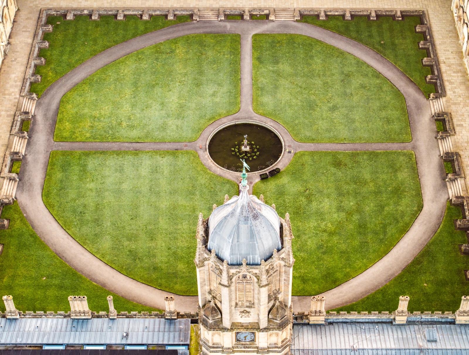 The dome of the Radcliffe Camera intrudes into the lower frame as a drone directly above traces the formal geometry of Radcliffe Square, Oxford. Circular gravel paths divide four quadrants of close-mown lawn, converging on a central fountain pool. The flat March light suppresses shadow and reduces the scene to near-diagrammatic form.