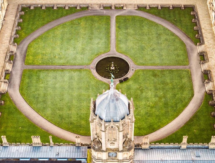 The dome of the Radcliffe Camera intrudes into the lower frame as a drone directly above traces the formal geometry of Radcliffe Square, Oxford. Circular gravel paths divide four quadrants of close-mown lawn, converging on a central fountain pool. The flat March light suppresses shadow and reduces the scene to near-diagrammatic form.