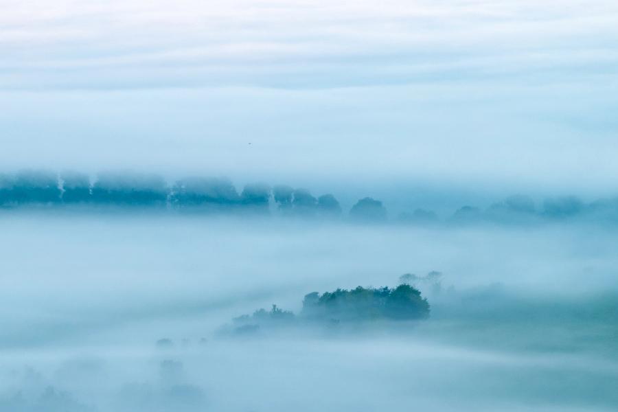A temperature inversion settles over the Thames Valley near Henley, reducing the lowland to layered planes of pale blue fog. Isolated tree canopies emerge as dark islands, the furthest treeline fading almost entirely into the overcast sky. The graduated recession of tones gives the scene an almost monochromatic, painterly quietness.