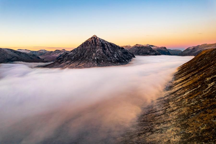 A temperature inversion fills Glen Etive and Glencoe at dawn, leaving Buachaille Etive Beag isolated above a smooth sea of cloud. Shot from an elevated drone position above the eastern ridge, the pyramid peak catches the first warm light against a sky still holding the blue of night. The foreground slope anchors the composition, giving scale to the suspended cloudscape below.