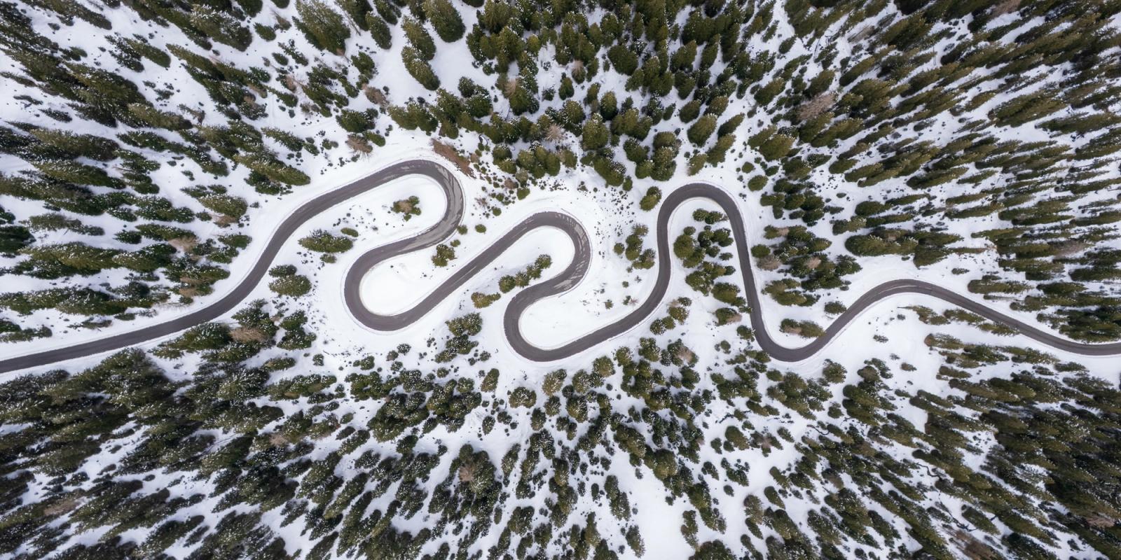 A serpentine mountain pass cuts through a snow-covered conifer forest above Cortina d'Ampezzo, photographed directly overhead at midday. The cleared asphalt road forms a continuous sinuous line across the white ground plane, its curves repeated with near-graphic regularity. Snow between the trees flattens the scene into a high-contrast pattern of grey, white, and dark green.