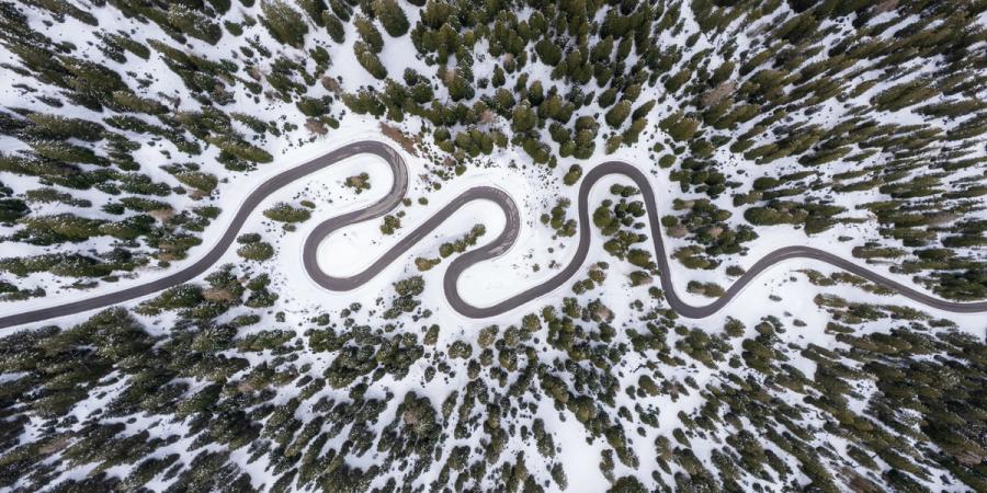 A serpentine mountain pass cuts through a snow-covered conifer forest above Cortina d'Ampezzo, photographed directly overhead at midday. The cleared asphalt road forms a continuous sinuous line across the white ground plane, its curves repeated with near-graphic regularity. Snow between the trees flattens the scene into a high-contrast pattern of grey, white, and dark green.