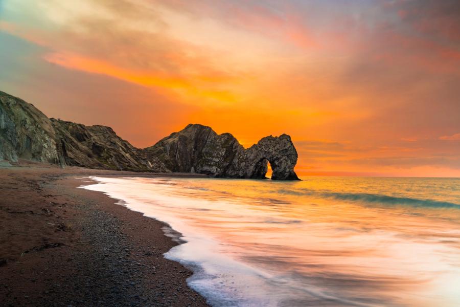 Durdle Door at sunset on a January evening, the long exposure smoothing the incoming tide into pale silk against the shingle. The natural limestone arch frames the burning horizon, its silhouette anchoring a sky of deep amber and rose.