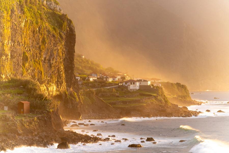Warm evening light rakes across a moss-covered sea cliff on Madeira's north coast, catching spray-haze above the shoreline and illuminating a small terraced settlement clinging to the headland. Breaking Atlantic swell scatters around dark basalt rocks in the foreground, while a storm sky above deepens the contrast with the golden low sun. The atmospheric haze gives the scene an unusual luminosity that lifts it beyond straightforward travel record.