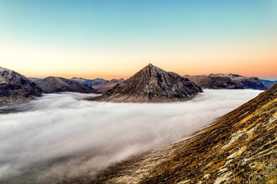 A pyramidal peak rises clear of a temperature inversion that has filled the surrounding glens with cloud, photographed from altitude at dawn in mid-winter. The peach-toned afterglow along the horizon separates the cloud sea from a cool blue sky, compressing the scene into three distinct horizontal bands. The composition is strengthened by the rocky foreground ridge leading the eye toward the isolated summit.