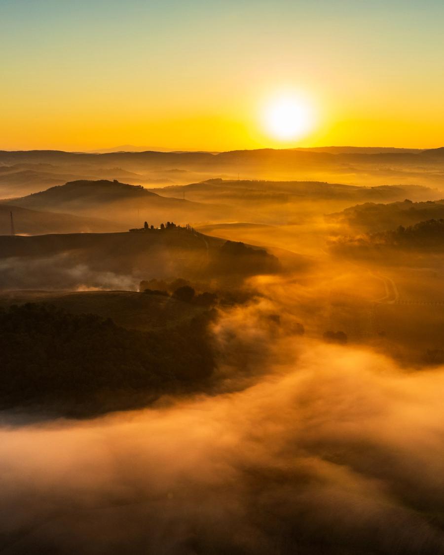 Dawn mist pools in the valleys of the Crete Senesi, leaving isolated hilltop farms and cypress lines as dark silhouettes against the rising sun. A temperature inversion holds the fog in the hollows, creating layered planes that recede toward the horizon. Captured by drone at first light on a still September morning.