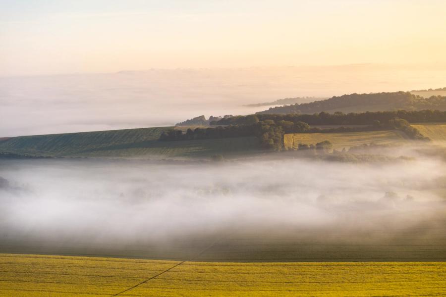 A temperature inversion fills the valley below the Chiltern escarpment near Henley-on-Thames, the fog pooling in horizontal bands while the higher groundâarable fields, mixed woodland, and a distant treelineâemerges in early morning warmth. The layering of mist, hill, and pale sky creates a compressed, almost tonal recession that rewards careful looking. Captured from altitude, the aerial perspective clarifies the topography in a way ground-level shooting could not.