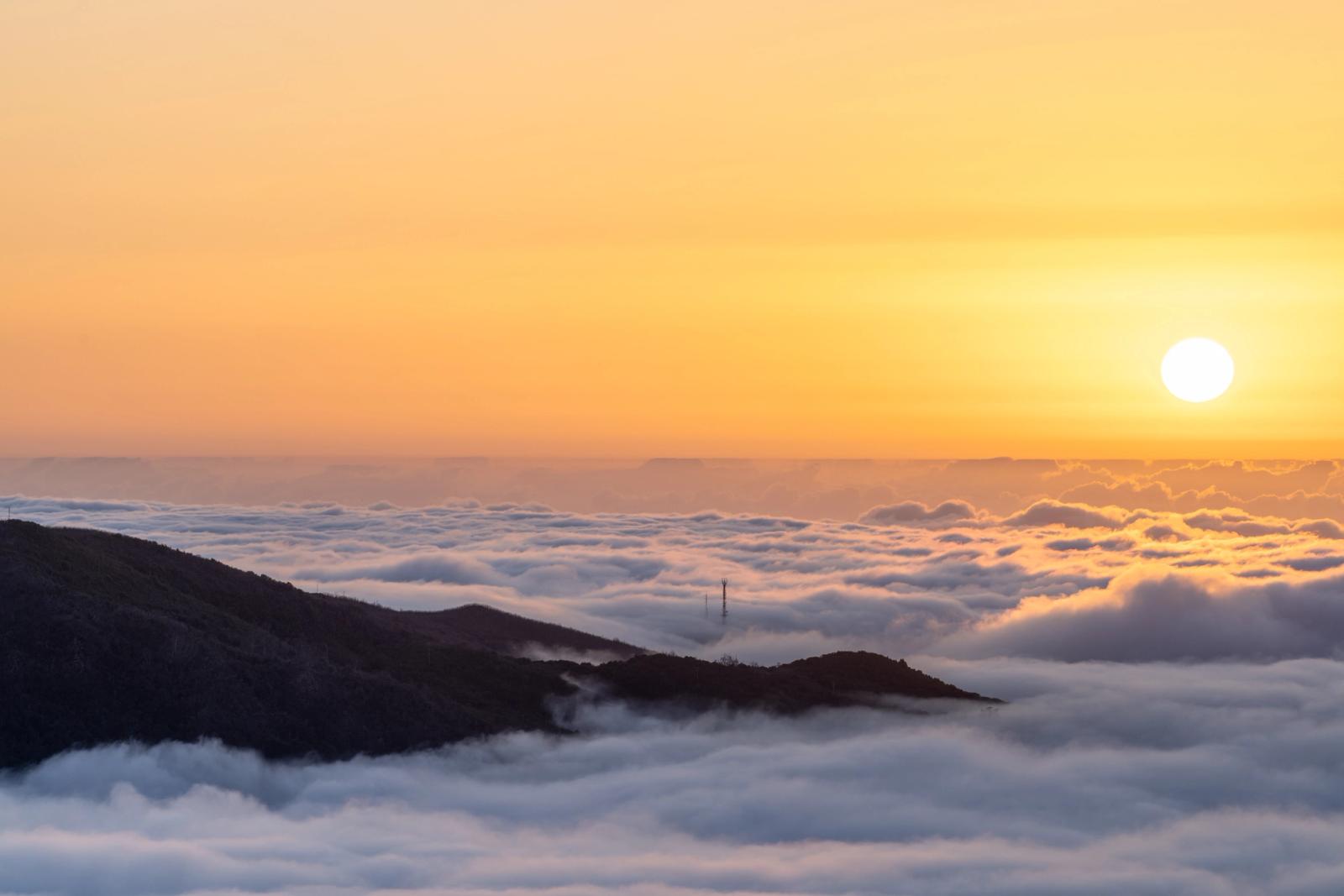 A temperature inversion fills the valleys below a mountain ridge, the cloud layer stretching unbroken to the horizon as the sun sits low and unobscured in a clear amber sky. A communications mast protrudes just above the cloud surface, a small note of the human world otherwise submerged. The image is taken from a high summit vantage at dusk or late evening in late May.