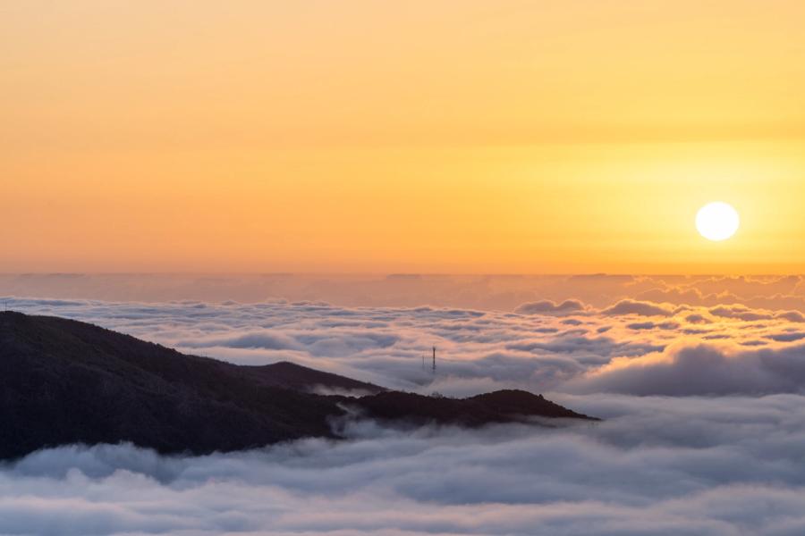 A temperature inversion fills the valleys below a mountain ridge, the cloud layer stretching unbroken to the horizon as the sun sits low and unobscured in a clear amber sky. A communications mast protrudes just above the cloud surface, a small note of the human world otherwise submerged. The image is taken from a high summit vantage at dusk or late evening in late May.