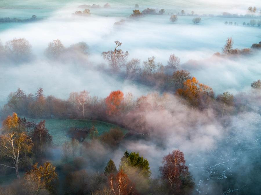 A temperature inversion settles across the Thames Valley near Wallingford, submerging hedgerows and fields beneath a continuous white plane while isolated trees in late autumn / early winter colour rise through the fog layer. Shot at low drone altitude, the diagonal ridge of emerging woodland divides the composition, giving structure to an otherwise formless sea of mist. The pale warmth catching copper and amber crowns against the cool tonal frozen ground is what lifts this from record to image.