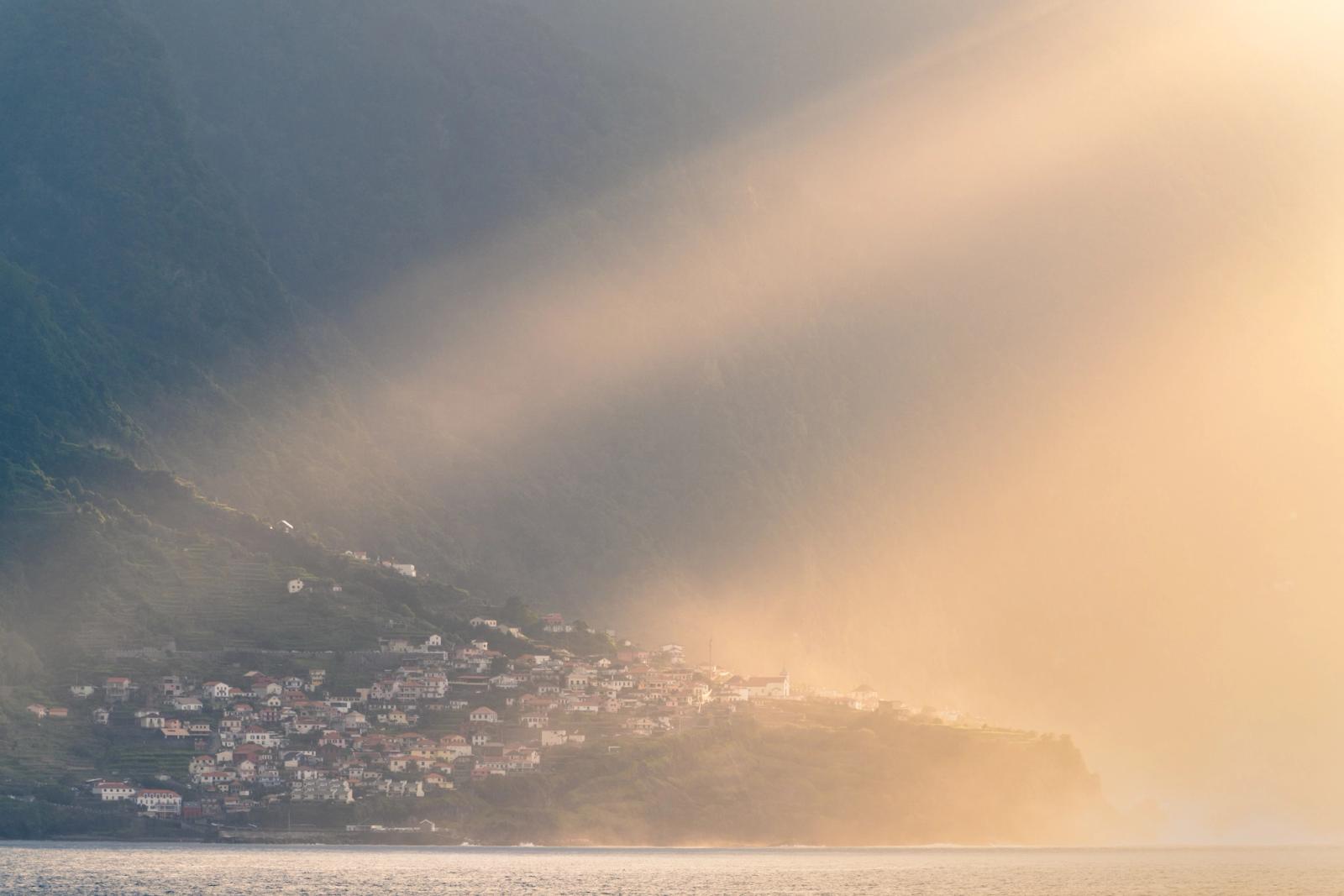 A coastal settlement clings to steep terraced slopes as a wall of mist rolls in from the sea, splitting the scene between cool shadow and warm evening light. The village â most likely on Madeira's north coast â is caught at the threshold of erasure, the golden haze reducing white-walled houses to silhouette at its margin.