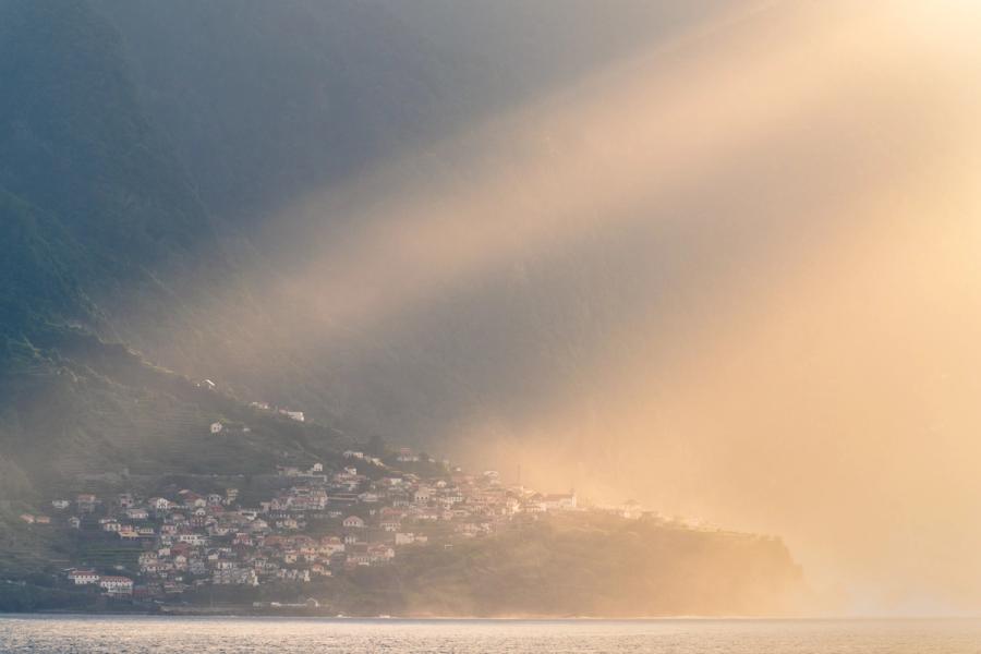 A coastal settlement clings to steep terraced slopes as a wall of mist rolls in from the sea, splitting the scene between cool shadow and warm evening light. The village â most likely on Madeira's north coast â is caught at the threshold of erasure, the golden haze reducing white-walled houses to silhouette at its margin.
