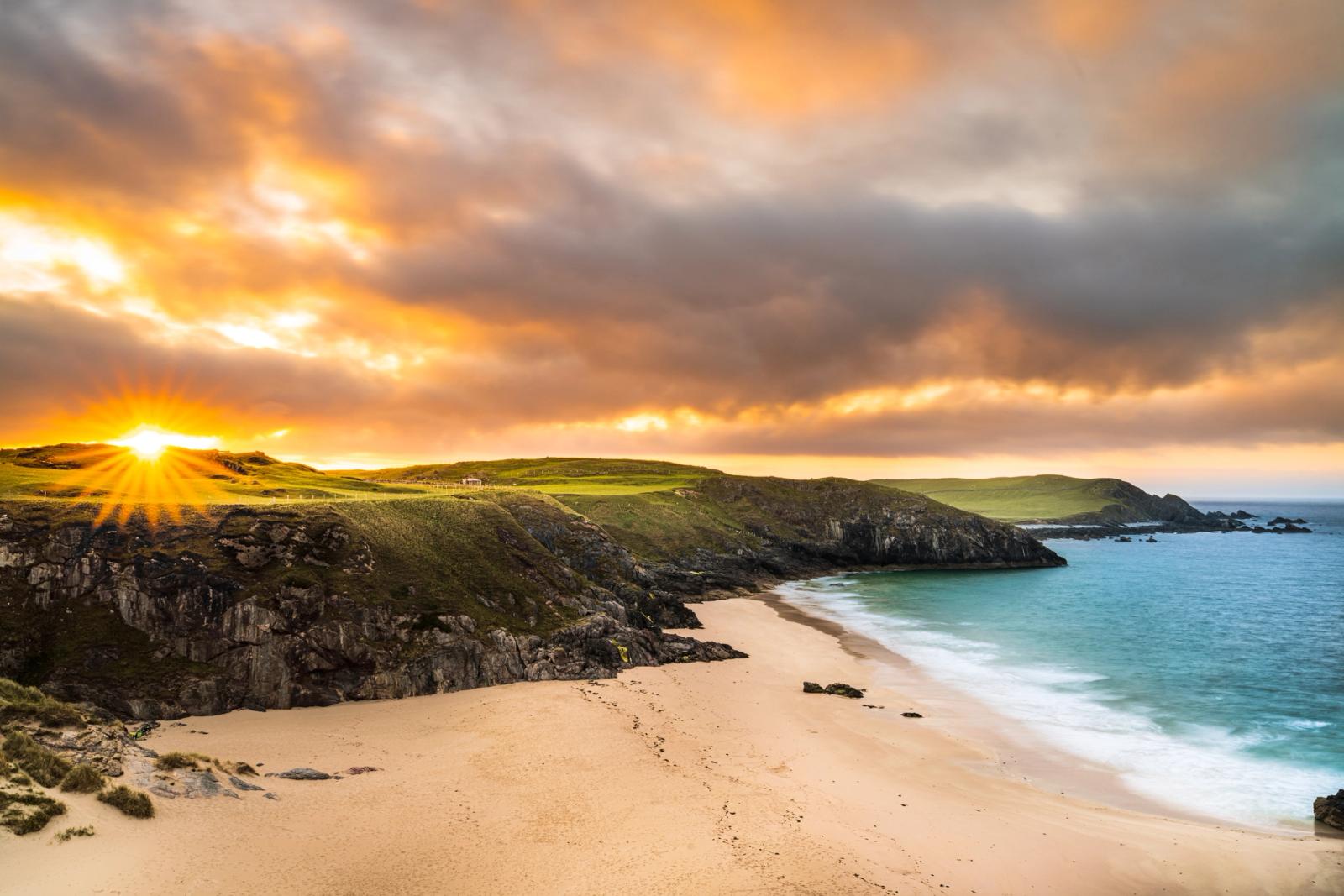 A low sun breaks through storm cloud above a horseshoe bay, casting starburst rays across green clifftop turf and pale sand. The long exposure smooths the incoming Atlantic swell to a pale wash against the beach, while darker cumulus holds the eastern sky. The cliffs, beach geometry, and quality of Atlantic light suggest the north-west Scottish coast.