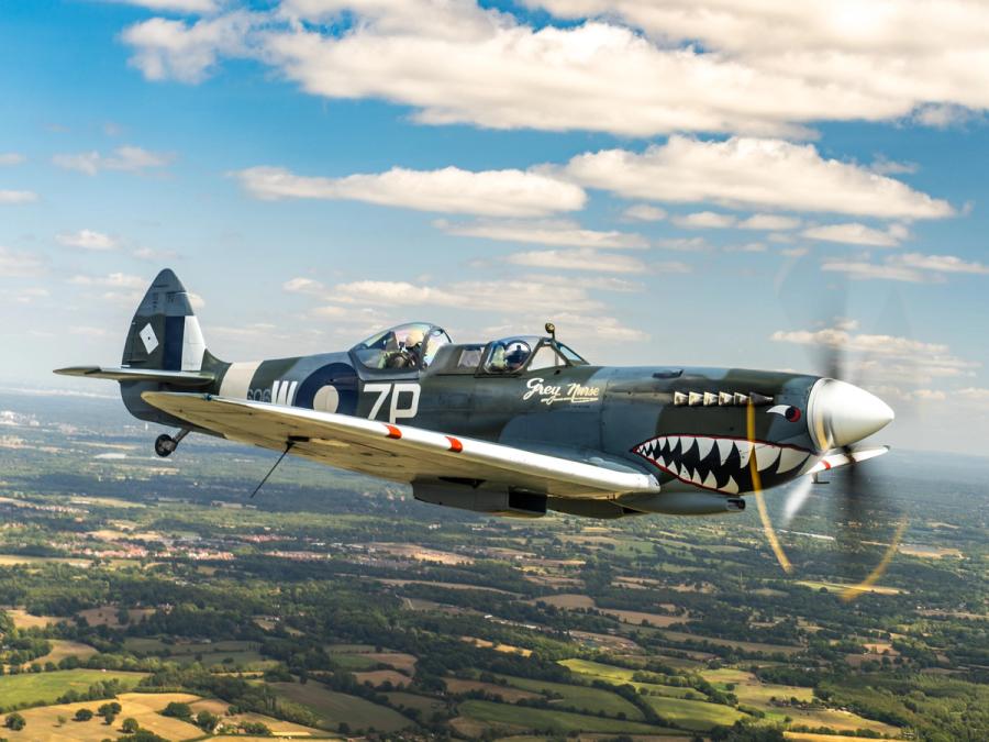 A two-seat Supermarine Spitfire trainer finished in shark-mouth livery and marked 'Grey Nurse' crosses above a patchwork of summer fields and woodland. The spinning propeller blurs against a partial cumulus sky, placing the aircraft in mid-flight isolation. The image documents an air-to-air encounter rather than a landscape moment.