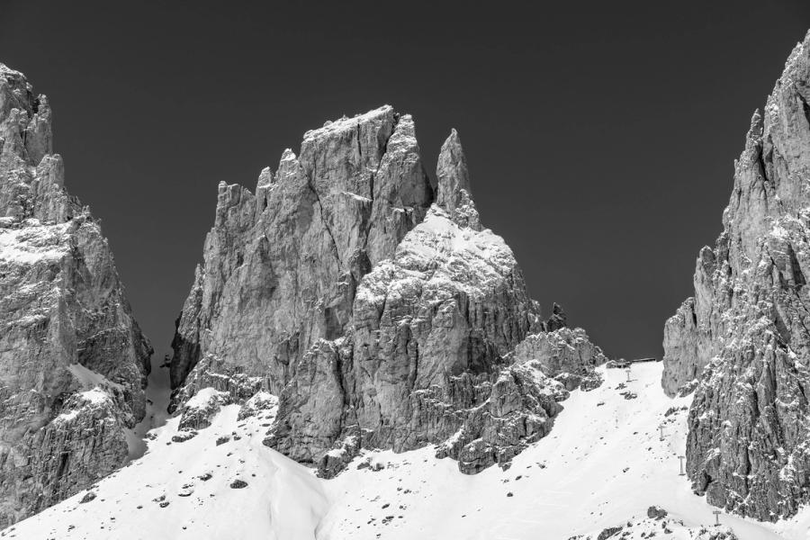 Jagged limestone towers rise from deep winter snowfields, their fractured faces rendered in hard light against a darkened sky. The monochrome conversion amplifies the tonal contrast between sun-struck rock and shadow, giving the spires a graphic, almost architectural weight. A ski lift visible at the right edge anchors the scene in managed mountain terrain.