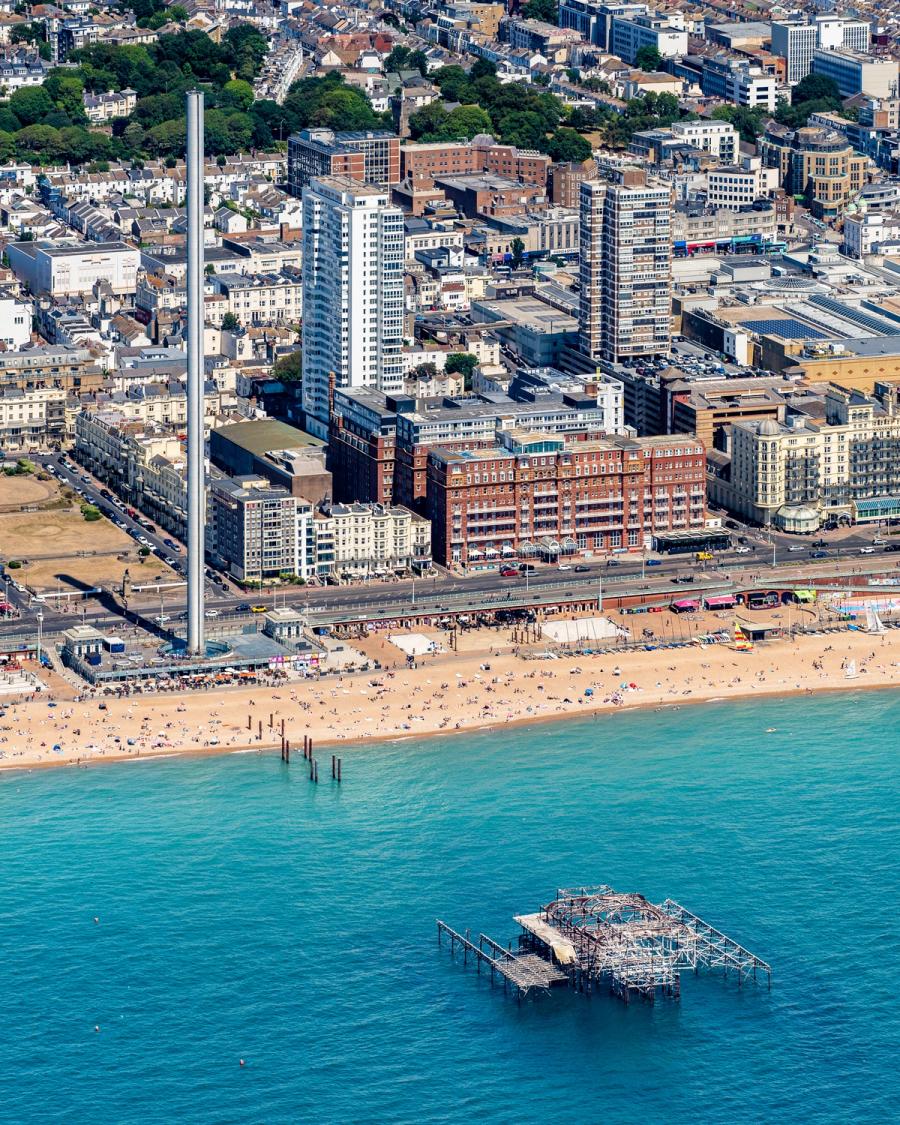 The skeletal remains of Brighton's West Pier sit isolated in the English Channel, framed against a crowded summer beach and the dense Victorian and modern townscape beyond. Shot from a high vantage point in strong midday light, the image documents rather than interprets, with the ruined iron structure providing the sole focal anchor in an otherwise busy frame.
