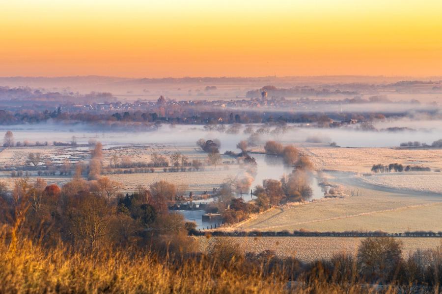 A temperature inversion holds low mist across a frost-silvered floodplain at first light, a river lock and its attendant willows emerging from the white. The amber pre-sunrise sky grades into the haze, compressing the middle distance into soft receding planes. A distant market town, barely resolved through the mist, anchors the human scale against the stillness.