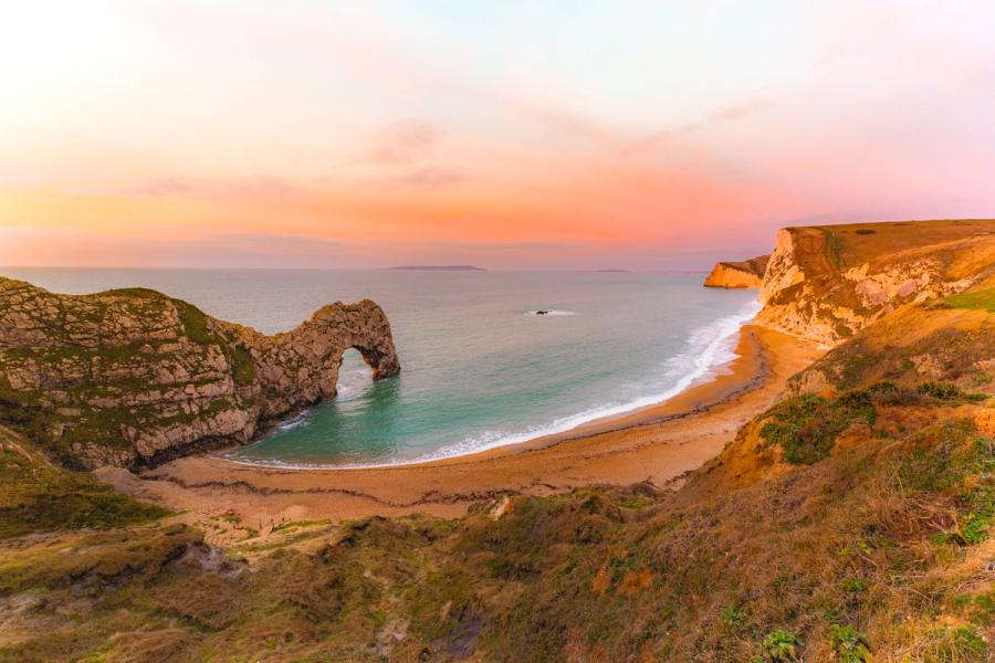 Durdle Door at last light in late January, the limestone arch silhouetted against a graduated pink and amber sky. The elevated viewpoint encompasses the full sweep of the bay, warm-toned cliffs framing turquoise shallows. A distant island sits on the horizon beneath receding cloud.