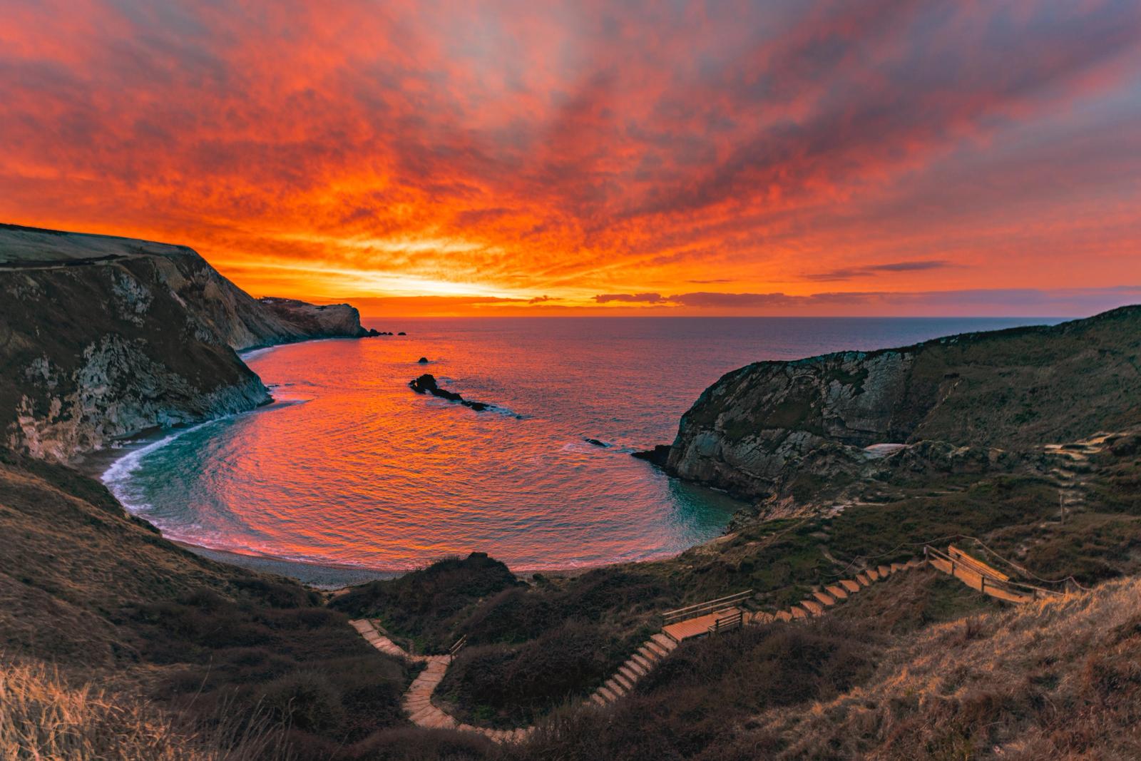 A sheltered coastal cove opens to the sea beneath a sky ignited at sunset, the calm water mirroring orange and crimson cloud. A stepped path and wooden railing descend from the clifftop foreground, anchoring the composition in the human scale of the landscape. Captured in late January, the low winter sun sits just below the horizon, its afterglow saturating both sky and sea surface.