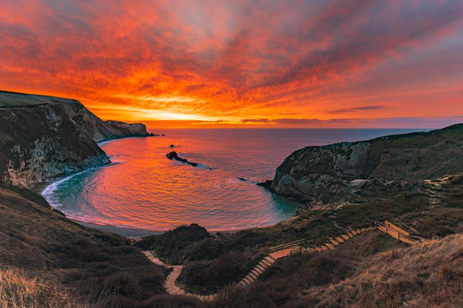 A sheltered coastal cove opens to the sea beneath a sky ignited at sunset, the calm water mirroring orange and crimson cloud. A stepped path and wooden railing descend from the clifftop foreground, anchoring the composition in the human scale of the landscape. Captured in late January, the low winter sun sits just below the horizon, its afterglow saturating both sky and sea surface.