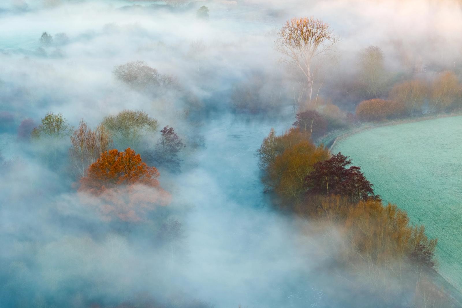 A temperature inversion smothers the Thames valley near Abingdon at first light, leaving trees in full autumn-to-winter transition stranded above a slow-moving sea of cold fog. A frosted meadow breaks clear to the right, its pale green a quiet counterpoint to the amber and burgundy crowns emerging from the mist. The aerial perspective dissolves conventional landscape geometry, reducing the scene to colour, texture, and atmosphere.