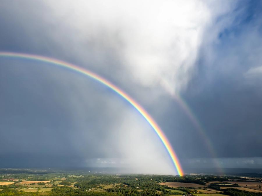 A double rainbow descends through a breaking storm cell over the pastoral patchwork south of Henley-on-Thames, the inner arc saturated against a dark cumulonimbus curtain while virga trails beneath the cloud base. The elevated drone viewpoint compresses arc and storm into a single geometrically forceful frame. Captured late afternoon in the aftermath of a convective summer storm.