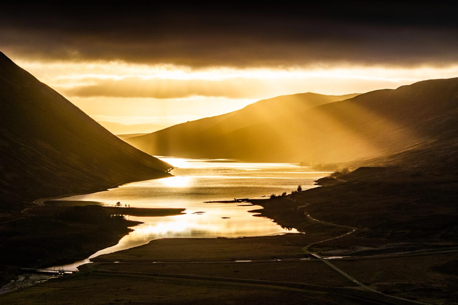 Crepuscular rays break through storm cloud above a Scottish loch near Pitlochry, casting raking winter light across still water and silhouetted hill flanks. The elevated viewpoint compresses the valley, aligning the loch's surface reflections with the source light in a single receding axis. Shot on Christmas Eve at low solar elevation, the geometry of cloud, mountain and water is unusually resolved.