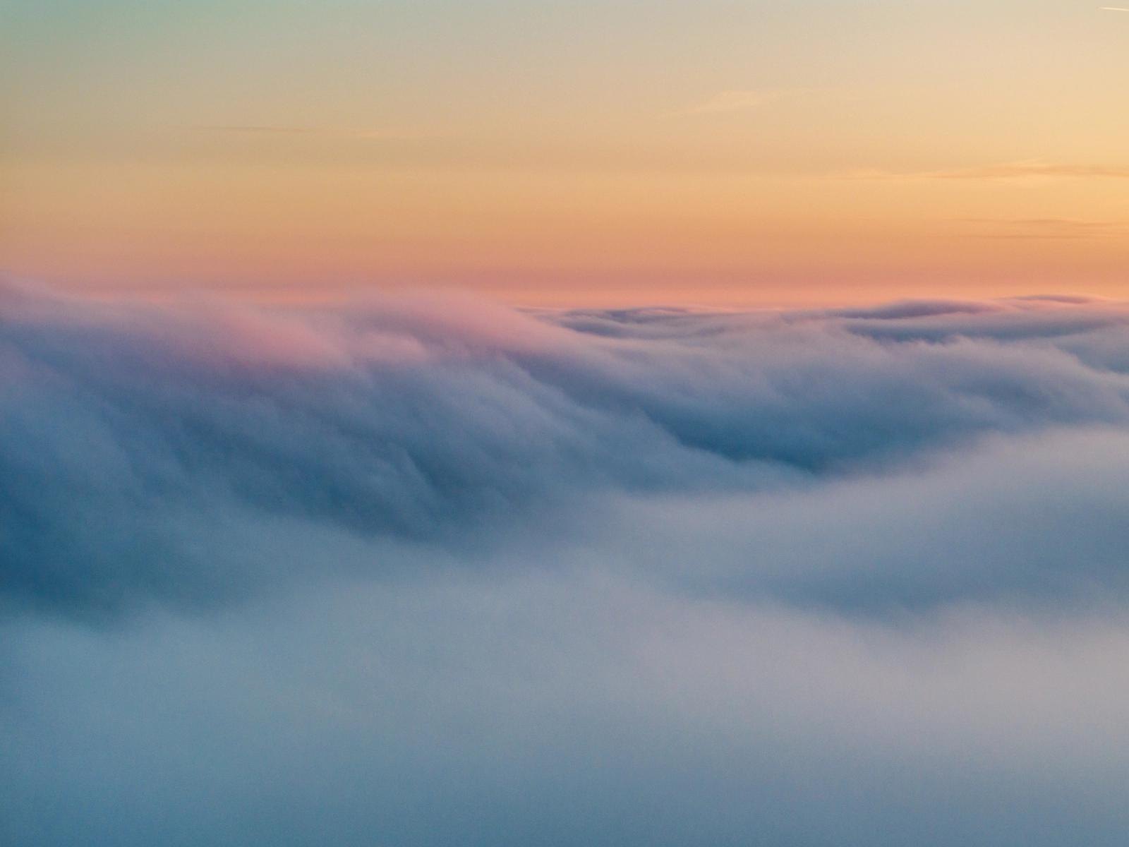 A temperature inversion buries the Thames Valley beneath a rolling sea of low cloud near Abingdon-on-Thames, the upper surface catching the last warm tones of a March sunset. Striations across the cloud top suggest gentle airflow, giving the otherwise featureless mass a quiet structural rhythm. The gradient from cool blue below to peach and teal at the horizon holds the eye without a conventional anchor point.