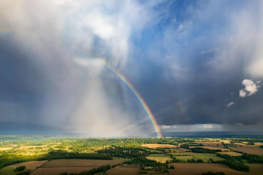 A vivid double rainbow descends through the base of a convective storm cell over the Thames Valley lowlands, captured from altitude as a rain shaft sweeps across a patchwork of summer fields and woodland. The aerial vantage reveals the full arc geometry impossible from ground level, set against a bruised, layered sky still broken by clear blue to the east. The proximity of sunlit crops and dark storm cloud gives the image an unusual tonal compression.
