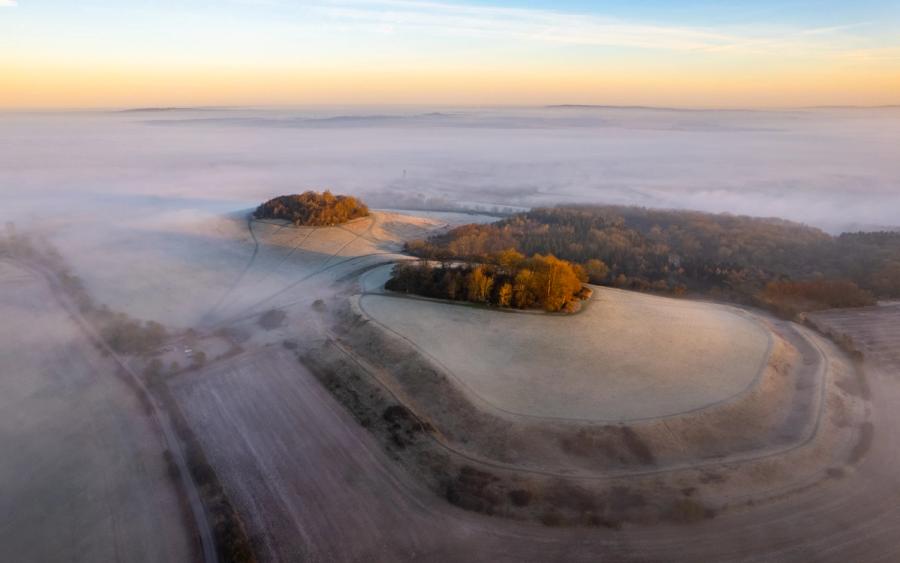 A temperature inversion buries the Vale of White Horse beneath a still sea of fog, viewed from Brightwell Barrow near the Wittenham Clumps in Oxfordshire, leaving frost-covered hilltops and copses of bare-branched trees standing clear in early dawn light. The curving field margins and converging ridge lines give the elevated ground an almost archipelago quality. Warm tones from the low sun catch the crowns of the remaining autumn-holdover foliage against the cold grey below.