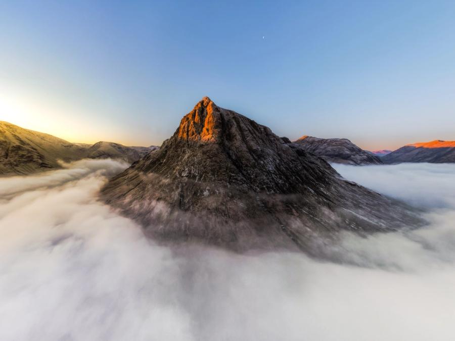 A temperature inversion buries the glens beneath the Glencoe Buachaille in a still sea of cloud, leaving the rocky summit emerging into first light above. The crescent moon holds its position in a steel-blue sky as alpenglow catches the peak's upper ridgeline. The surrounding peaks glow red from the sunrise.