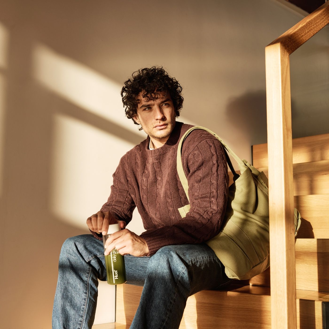 A man with curly hair sits on a wooden staircase, holding a bottle of Huel Daily Greens. He is dressed in a brown knit sweater, blue jeans, and has a green tote bag over his shoulder. Sunlight streams through a window, creating a warm, cozy atmosphere.