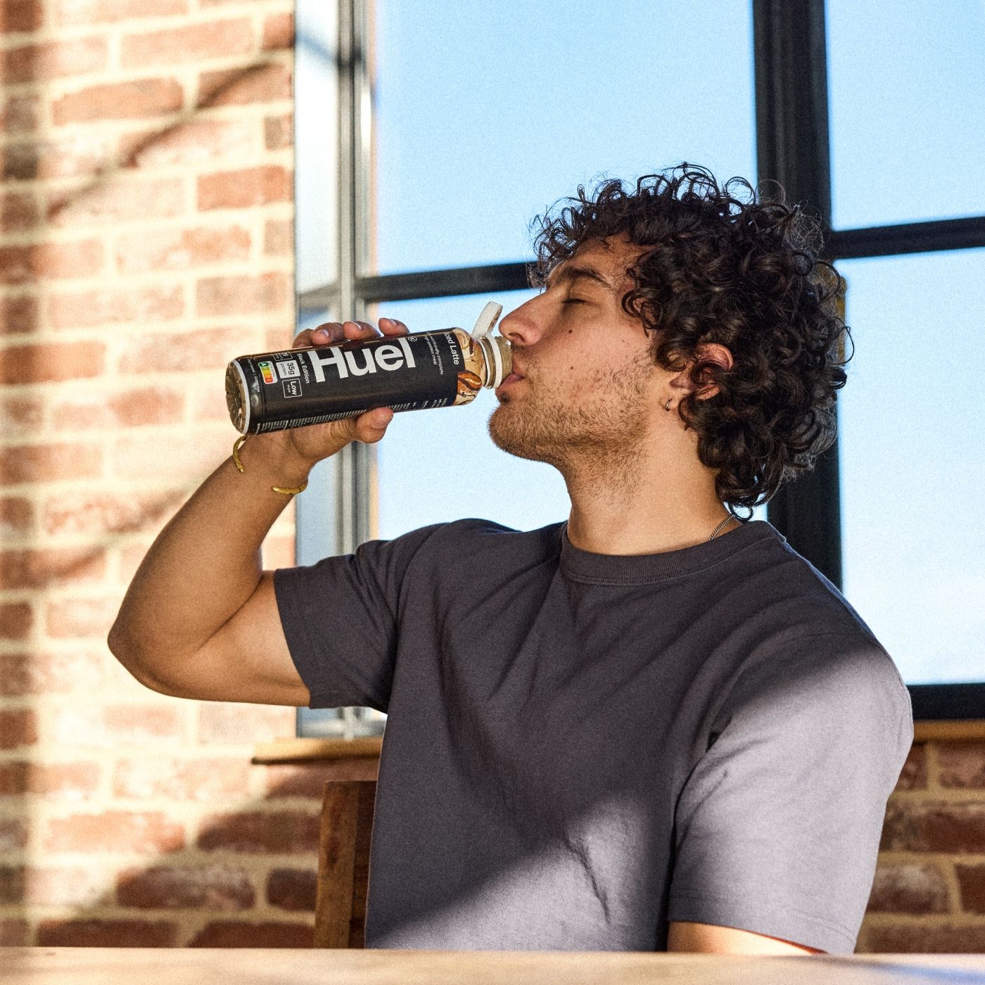 A man with curly hair is seated indoors, sipping from a chocolate-flavored Huel Ready-to-Drink bottle. He sits near a wooden table, with large windows and exposed brick walls in the background, creating a cozy and sunlit setting.