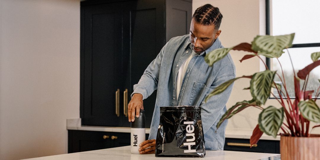 A man putting the lid on a black and white Huel shaker in a modern kitchen, next to a bag of black Huel powder