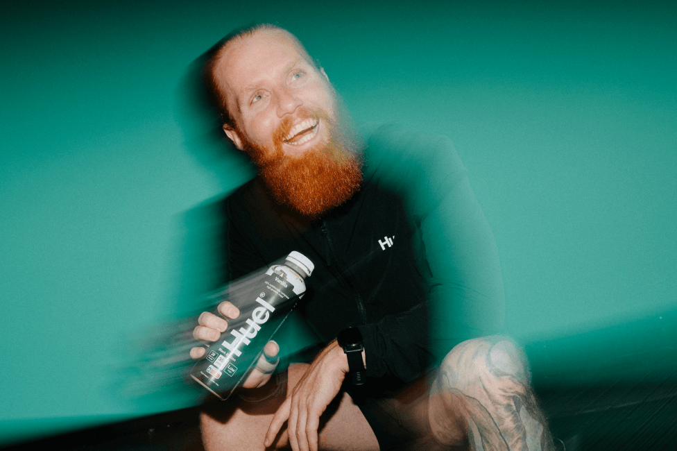 Endurance runner Russ Cook, The Hardest Geezer, kneeling while holding a bottle of Huel Ready-to-Drink against a teal background