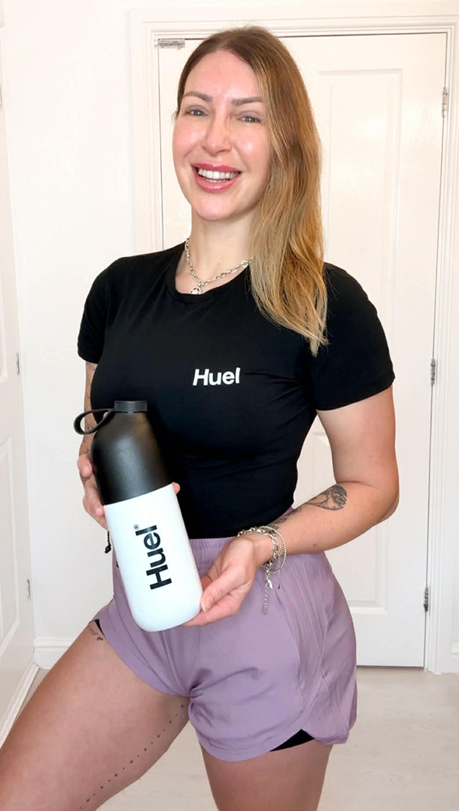 A lady wearing a black t-shirt and shorts smiling holding a Huel shaker