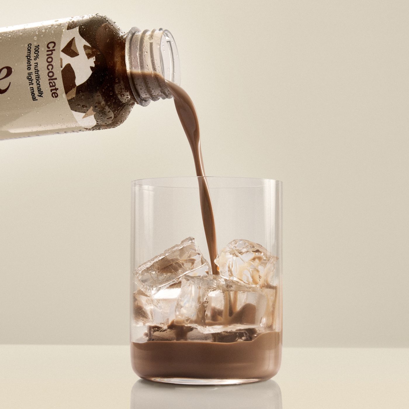 Huel chocolate pouring from a bottle of Huel Ready-to-drink lite into a glass with ice on a beige background