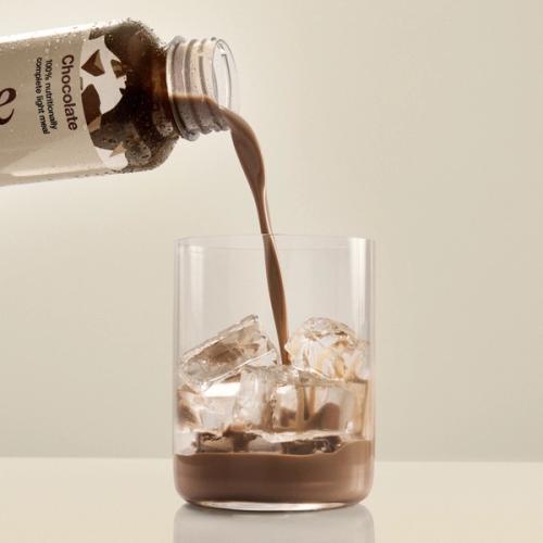 Huel chocolate pouring from a bottle of Huel Ready-to-drink lite into a glass with ice on a beige background