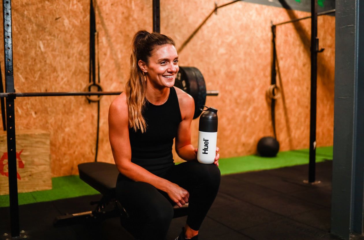 A Women smiling while sitting down in black gym clothes with gym equipment behind her resting a Huel Shaker on her knee