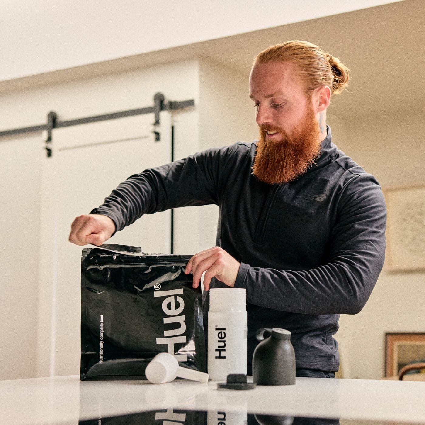 A man with a long red beard is standing in a modern kitchen, opening a large black bag of Huel Powder. On the counter in front of him are a white Huel shaker bottle and measuring scoop, suggesting he is preparing a meal replacement shake.