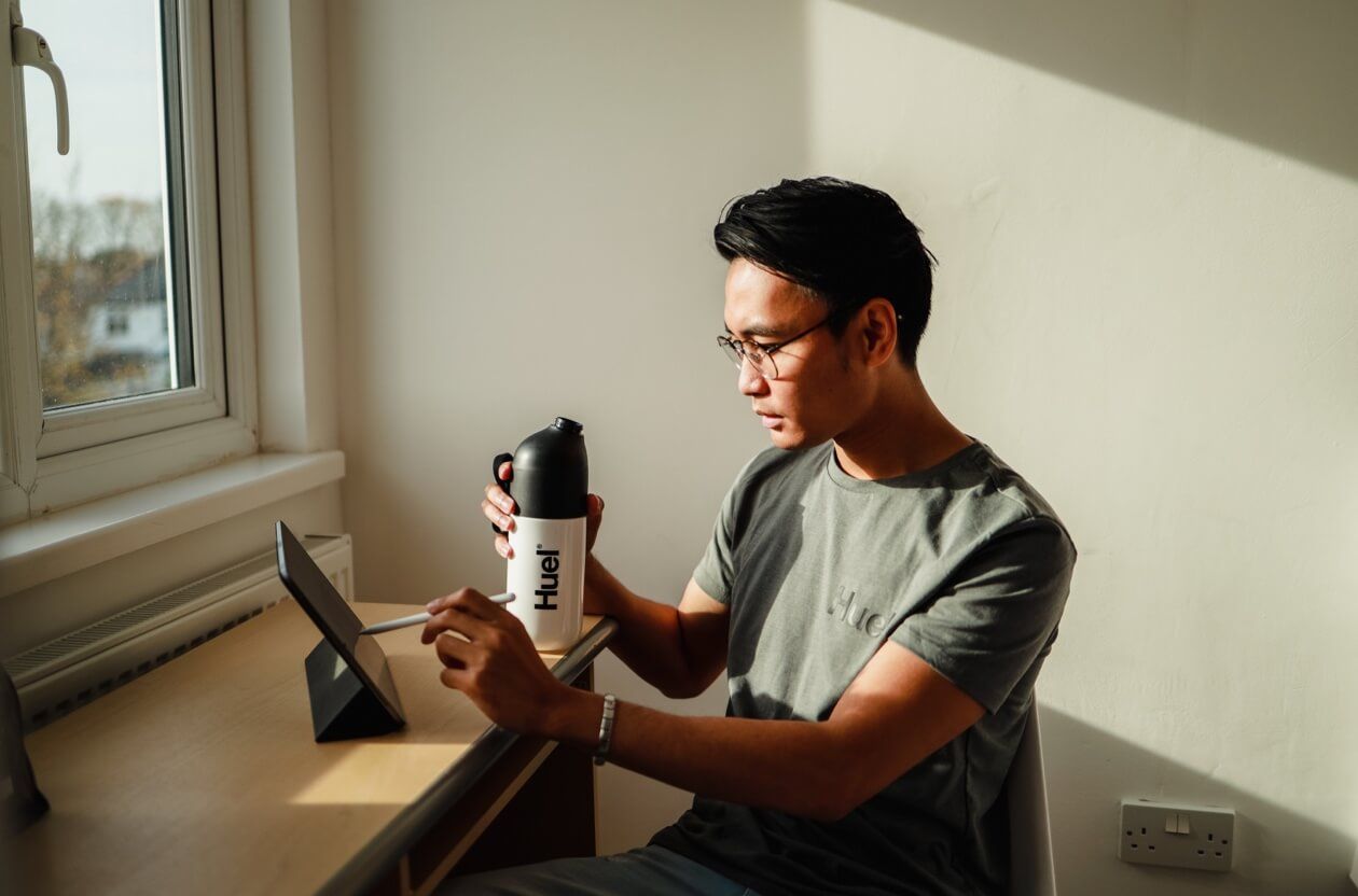 A Man in a grey Huel shirt sitting down at a desk in front of a window while holding a Huel shaker and working on a iPad